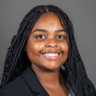 Studio portrait of a person with long braided hair, wearing a dark blazer and layered necklaces against a gray blue background. 
