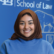 Person wearing a brown headscarf and a bright blue long-sleeve sweater with a black belt, standing indoors in front of a blue backdrop with the University at Buffalo School of Law logo and wooden panels. 