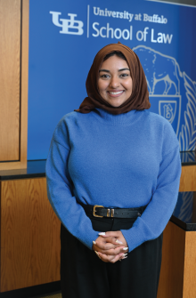 Person wearing a brown headscarf and a bright blue long-sleeve sweater with a black belt, standing indoors in front of a blue backdrop with the University at Buffalo School of Law logo and wooden panels. 
