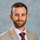 Studio portrait of a person wearing a tan suit jacket, white dress shirt, and patterned red tie against a textured blue green background. 