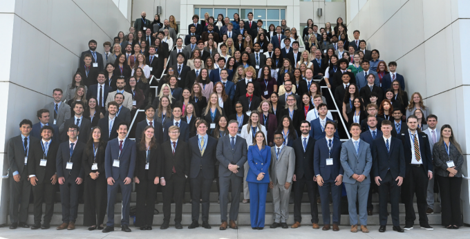 Large group photo of UB School of Law Class of 2028 posed on outdoor steps in front of a modern white building. Individuals are dressed in formal business attire, including suits, ties, and professional dresses, with name tags visible on many. 