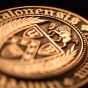 Close‑up of a bronze circular seal featuring a shield surrounded by a laurel wreath and encircled by raised Latin lettering. The seal is photographed at an angle against a dark background, with warm lighting highlighting the engraved details. 
