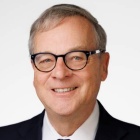 A head‑and‑shoulders professional portrait of David Franasiak wearing a dark suit jacket, white shirt, and dark tie, photographed against a light, plain background. The individual is wearing eyeglasses.