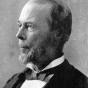 Black and white historical portrait of a person shown in side profile, wearing a dark formal jacket, white shirt, and bow tie. The person has neatly combed hair and a short beard, and is photographed against a plain studio background. 
