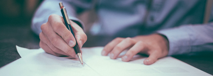 photo of a man holding a pen, writing on a piece of paper. 