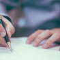 photo of a man holding a pen, writing on a piece of paper. 