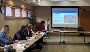 woman standing in a conference room in front of a large screen that is displaying a presentation. 