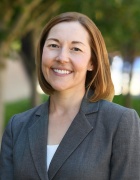 woman in a grey suit, standing outside in front of some trees. 