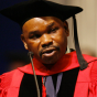 man wearing red graduation regalia, giving a speech. 