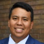 man smiling, wearing a blue business suit, standing in front of a brick wall. 