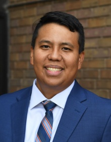 man smiling, wearing a blue business suit, standing in front of a brick wall. 