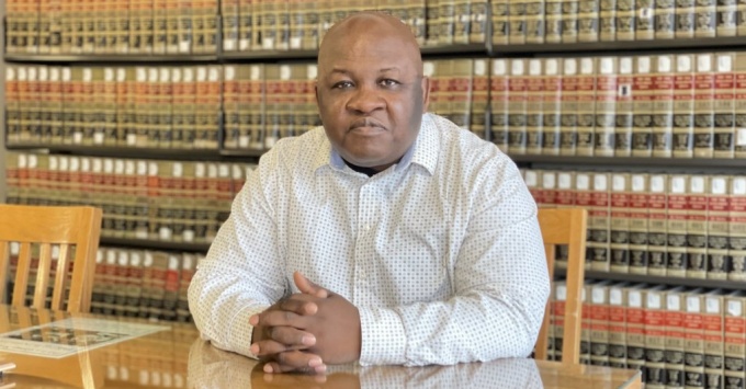 black gentleman wearing white shirt, sitting at a wooden table with shelved books behind him. 