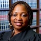 photo of a woman in judge's robe, holding a book, standing in front of library shelving. 