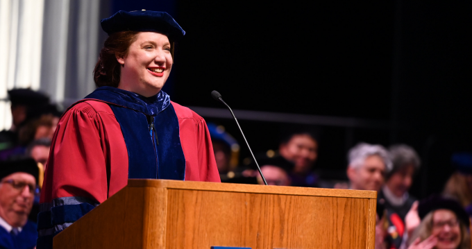 woman in red regalia standing at a podium, smiling. 