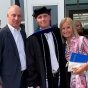 young man dressed in graduation regalia, standing between his mother and father outside a building. 