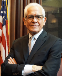 man wearing glasses, arms folded, standing in front of American flag. 