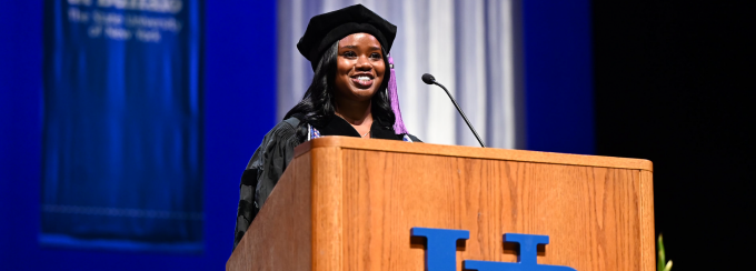 young woman wearing commencement regalia, standing at a podium. 