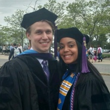 man and woman wearing graduation robes, standing outside, smiling. 