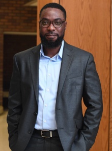 man wearing suit, glasses, standing inside a hallway, smiling. 