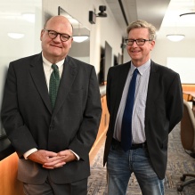 two men wearing glasses, standing next to a white board in a classroom. 