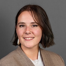 Young woman, smiling, wearing brown jacket, gold earrings. 