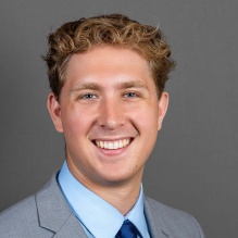 Young man, smiling, wearing blue/grey suit with blue tie. 