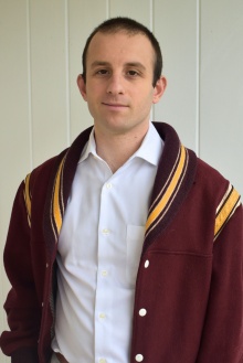 Young man wearing burgundy wool jacket, white button-down shirt, standing in a hallway. 