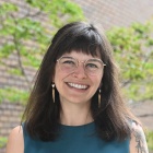 Woman wearing green dress, glasses, standing outside next to a brick buiding, smiling. 