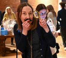Woman at an event, holding up fake mustache and magnifying glass. 