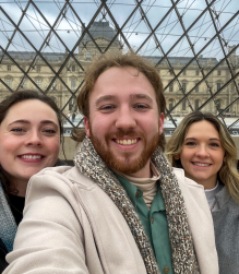 Three young adults, one male in the middle with two females, standing in front of a gate sepearating them from a large building in the background. 