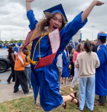 young woman, outside, wearing graduation regalia, jumping and smiling. 