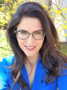 Woman wearing blue shirt, glasses, smiling, standing outside with tree branches behind her. 