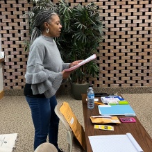 Woman reading aloud from a sheet of paper to a room of students. 