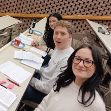 Three students sitting at a long table in a classroom, smiling. 
