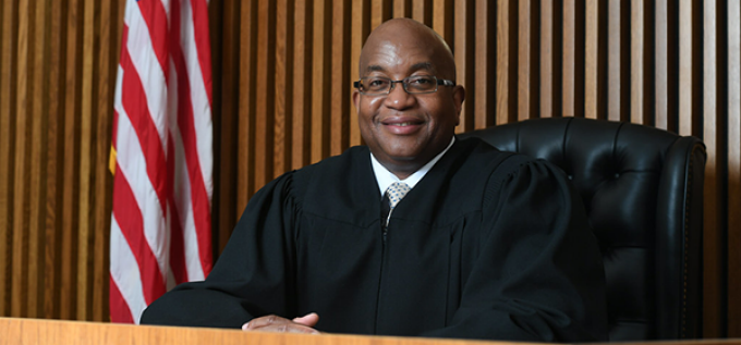 Smiling man wearing judges robe, sitting at a courtroom bench next to a U.S. flag. 