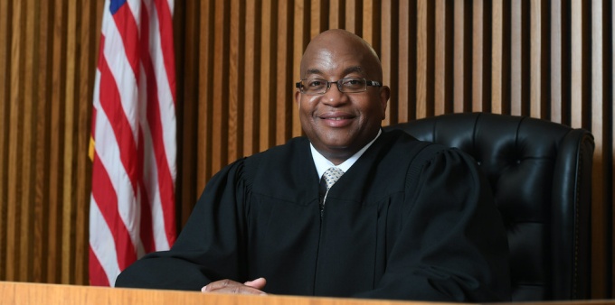 Smiling man wearing judges robe, sitting at a courtroom bench next to a U.S. flag. 