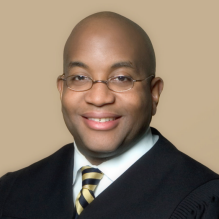 Man wearing black judges robe, tie, glasses, smiling. 