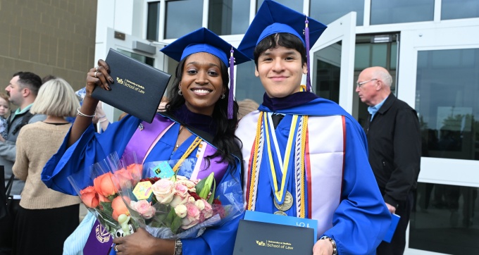 Young woman and man wearing graduation regalia, smiling, holding up diplomas. 