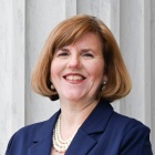 A person wearing a dark blue suit jacket over a light top with a pearl necklace, standing in front of large white marble columns. 