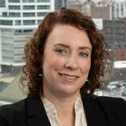 A person wearing a black blazer over a light blouse. Behind the desk is a large window showing a cityscape with multiple buildings. 