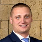 A person wearing a dark navy suit jacket, white dress shirt, and light blue necktie, standing with arms crossed in front of a tan stone wall with a rectangular block pattern. 