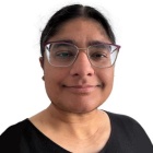 A person wearing a black top with a rounded neckline and glasses with dark-colored frames, shown against a plain white background. 