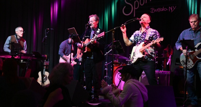 Live band performing on stage at Sportsmen’s Tavern in Buffalo, NY, with musicians playing guitars, keyboard, and drums under purple and green stage lighting. 