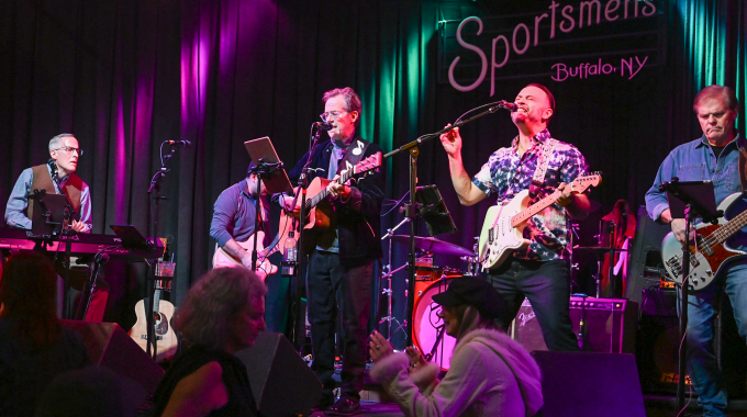 Live band performing on stage at Sportsmen’s Tavern in Buffalo, NY, with musicians playing guitars, keyboard, and drums under purple and green stage lighting. 