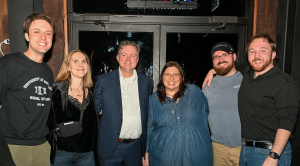 Group of six people standing together indoors in front of glass doors, dressed in casual and semi-formal clothing. 