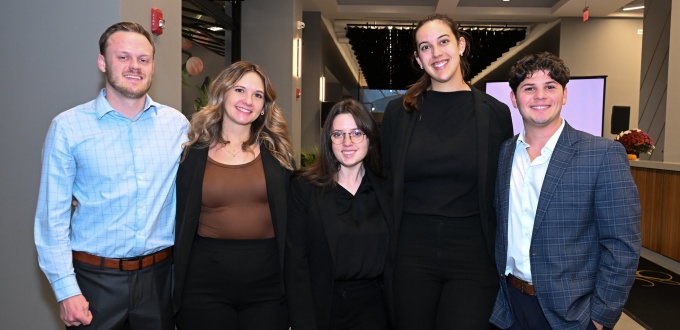 Five people standing together indoors in a modern, well-lit space with a high ceiling and decorative lights, dressed in business or semi-formal attire. 
