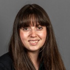 Person wearing a dark blazer over a green top, with long straight brown hair, posed against a plain gray background. 