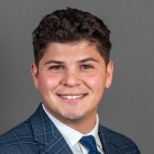 Person wearing a blue plaid suit jacket, white dress shirt, and solid blue tie against a plain gray background. 