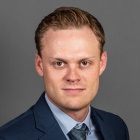 Person wearing a dark blue suit jacket, light blue dress shirt, and a textured gray tie, posed against a plain gray background. 