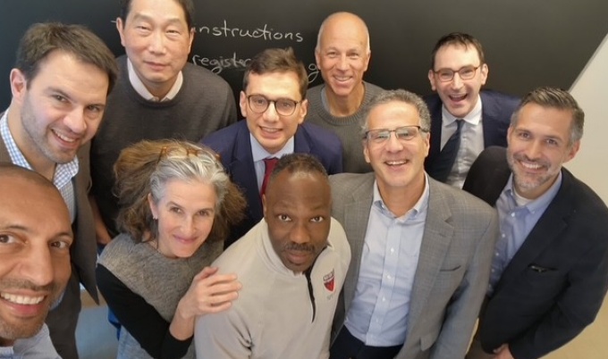 A group of people standing closely together in an indoor setting, posing for a photo taken from an elevated angle. They are dressed in business and casual attire, including blazers, sweaters, and collared shirts. Behind them is a blackboard with partially visible handwritten text. 